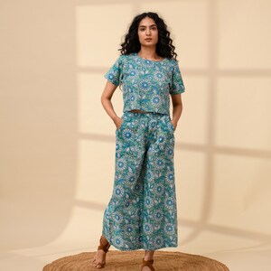 May include: A woman is wearing a blue and green floral print top and matching trousers. The top is a cropped style and the trousers are wide-legged. She is standing on a brown rug.