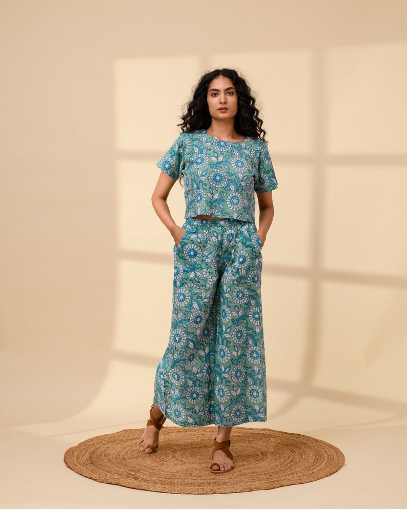 May include: A woman is wearing a blue and green floral print top and matching trousers. The top is a cropped style and the trousers are wide-legged. She is standing on a brown rug.