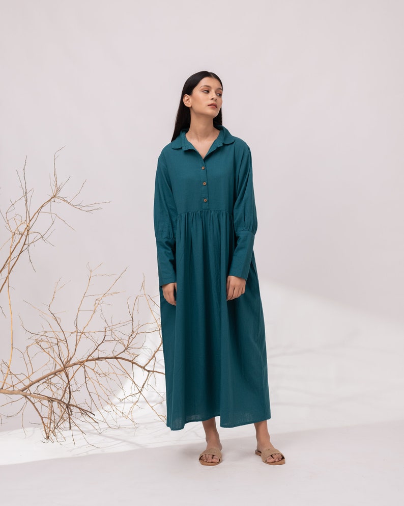 May include: A teal blue long-sleeved dress with a collared neckline and button closure. The dress has a gathered skirt and is made of a lightweight fabric.