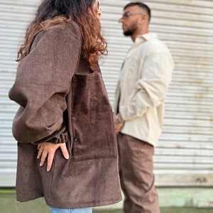 Puede incluir: Chaqueta de pana marr&oacute;n con cierre de bot&oacute;n y bolsillos grandes. La chaqueta se lleva con vaqueros azules. Una persona en el fondo lleva una camisa color crema y pantalones marrones.