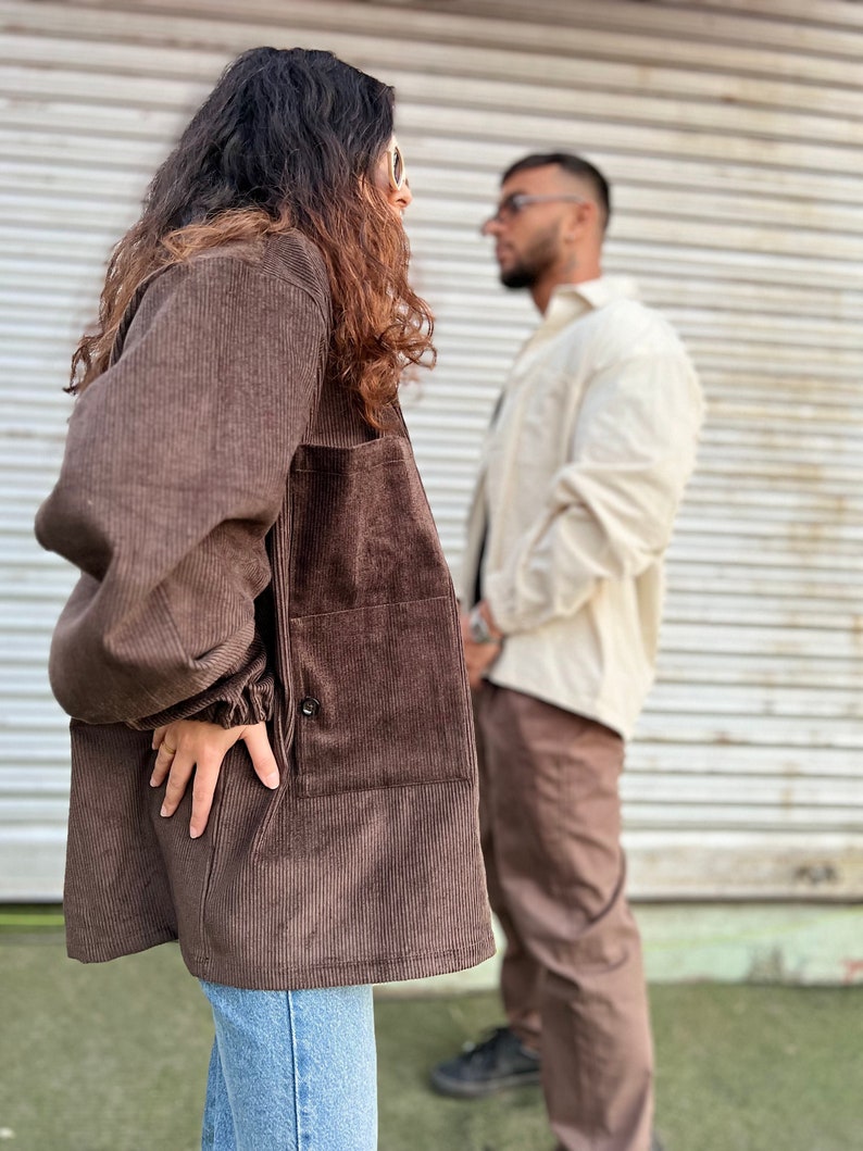 Puede incluir: Chaqueta de pana marr&oacute;n con cierre de bot&oacute;n y bolsillos grandes. La chaqueta se lleva con vaqueros azules. Una persona en el fondo lleva una camisa color crema y pantalones marrones.