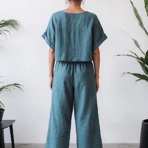 May include: Teal-colored linen two-piece outfit. The top is a short-sleeved, cropped shirt with a square neckline. The matching pants are wide-legged and cropped at the calf. The outfit is worn with black sandals.