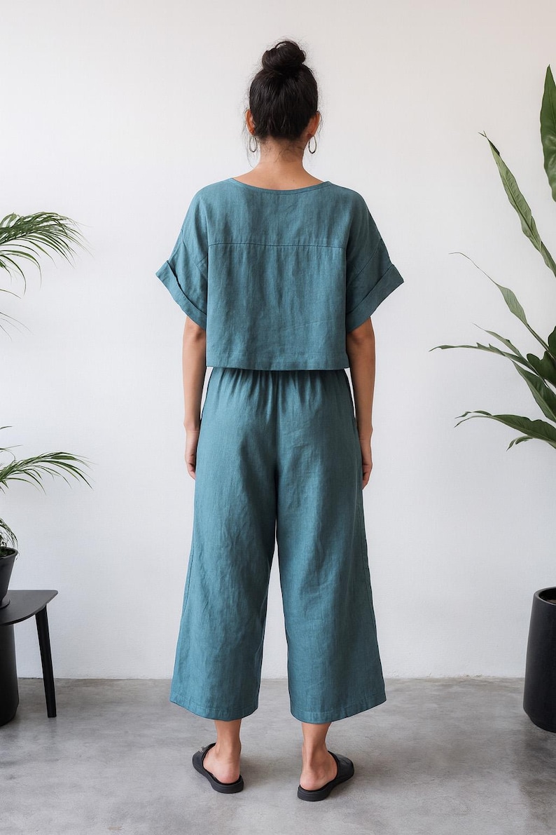 May include: Teal-colored linen two-piece outfit. The top is a short-sleeved, cropped shirt with a square neckline. The matching pants are wide-legged and cropped at the calf. The outfit is worn with black sandals.
