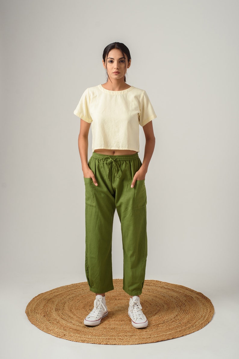 K&ouml;nnte beinhalten: Eine Frau tr&auml;gt ein hellgelbes, bauchfreies T-Shirt und eine olivgr&uuml;ne Hose mit Kordelzug. Sie steht auf einem runden gewebten Teppich und tr&auml;gt wei&szlig;e High-Top-Sneaker.