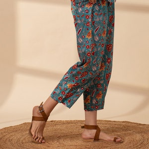 May include: Teal blue cropped trousers with a floral print in shades of red, orange, and white. The trousers have a wide leg and a relaxed fit.