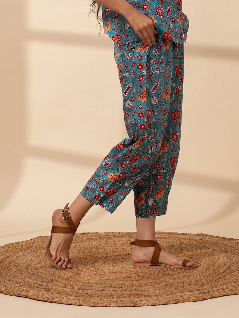 May include: Teal blue cropped trousers with a floral print in shades of red, orange, and white. The trousers have a wide leg and a relaxed fit.