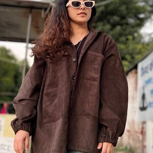 Puede incluir: Chaqueta de pana marr&oacute;n con cierre de botones, dos bolsillos delanteros y mangas largas. La chaqueta se lleva con una camisa negra y vaqueros azules. La persona lleva gafas de sol color crema.