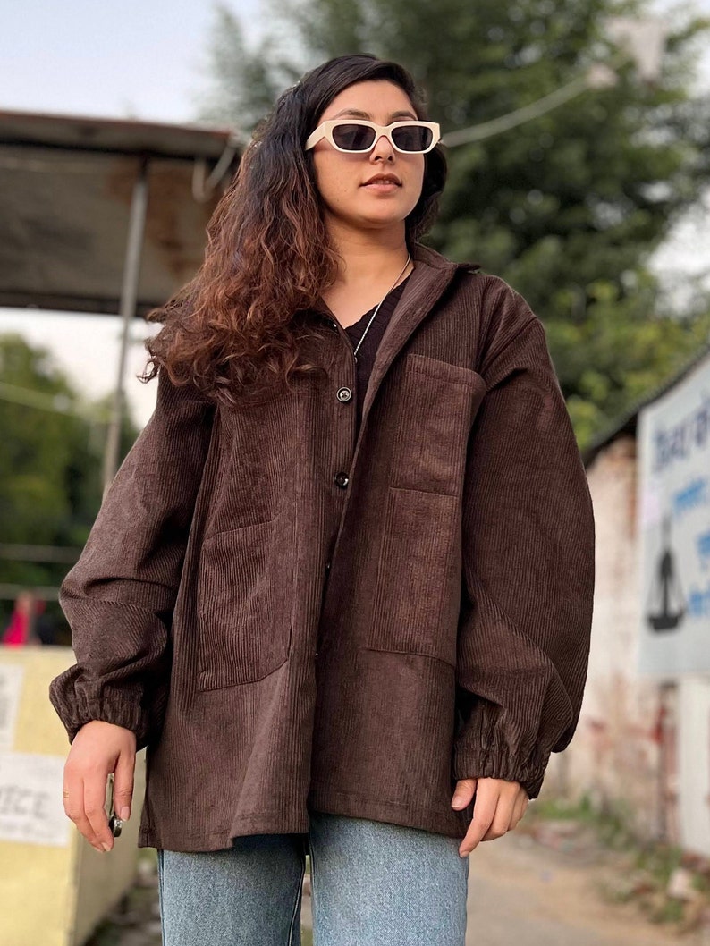 Puede incluir: Chaqueta de pana marr&oacute;n con cierre de botones, dos bolsillos delanteros y mangas largas. La chaqueta se lleva con una camisa negra y vaqueros azules. La persona lleva gafas de sol color crema.