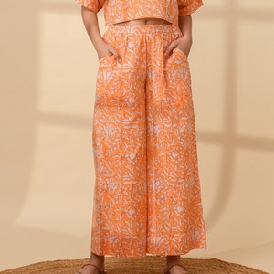 May include: An orange two-piece outfit with a floral pattern. The set includes a short-sleeved crop top and wide-leg pants with pockets. The outfit is worn with brown sandals on a woven mat.