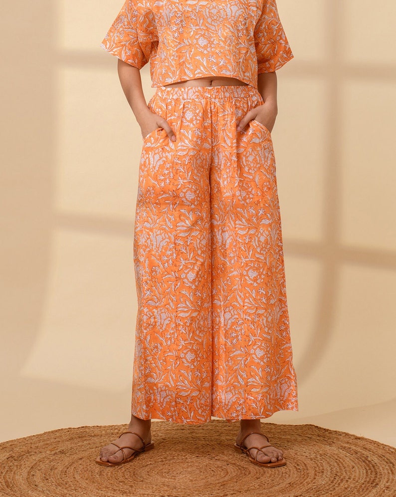 May include: An orange two-piece outfit with a floral pattern. The set includes a short-sleeved crop top and wide-leg pants with pockets. The outfit is worn with brown sandals on a woven mat.