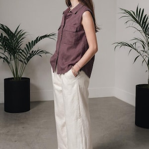 May include: A sleeveless, button-down shirt in a muted mauve color, paired with off-white wide-leg pants. The outfit is completed with black flip-flops. The setting is a minimalist interior with potted plants.
