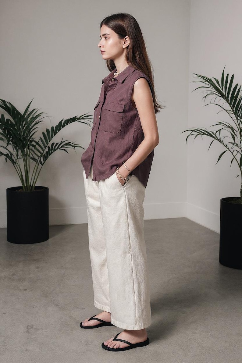 May include: A sleeveless, button-down shirt in a muted mauve color, paired with off-white wide-leg pants. The outfit is completed with black flip-flops. The setting is a minimalist interior with potted plants.