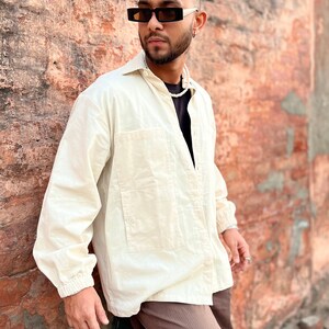 Puede incluir: Camisa crema con botones, dos bolsillos delanteros y mangas largas. La camisa se combina con pantalones marrones y gafas de sol negras. Un collar de perlas completa el atuendo. El modelo se apoya en una pared de ladrillo desgastada.