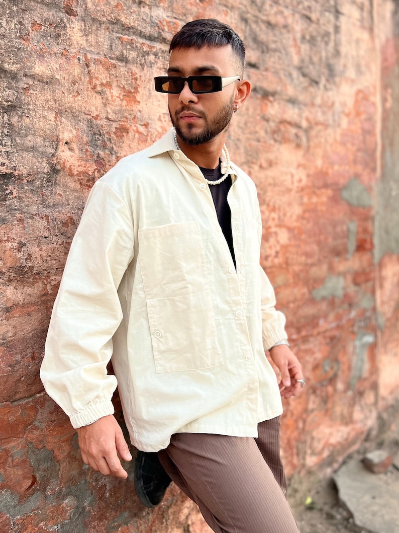 Puede incluir: Camisa crema con botones, dos bolsillos delanteros y mangas largas. La camisa se combina con pantalones marrones y gafas de sol negras. Un collar de perlas completa el atuendo. El modelo se apoya en una pared de ladrillo desgastada.