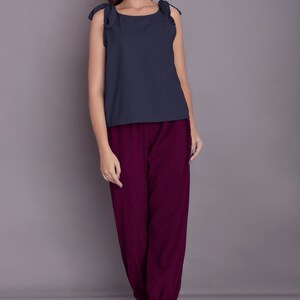 May include: A woman is wearing a dark blue sleeveless top with ruffled straps and burgundy trousers. She is standing on a light grey surface.