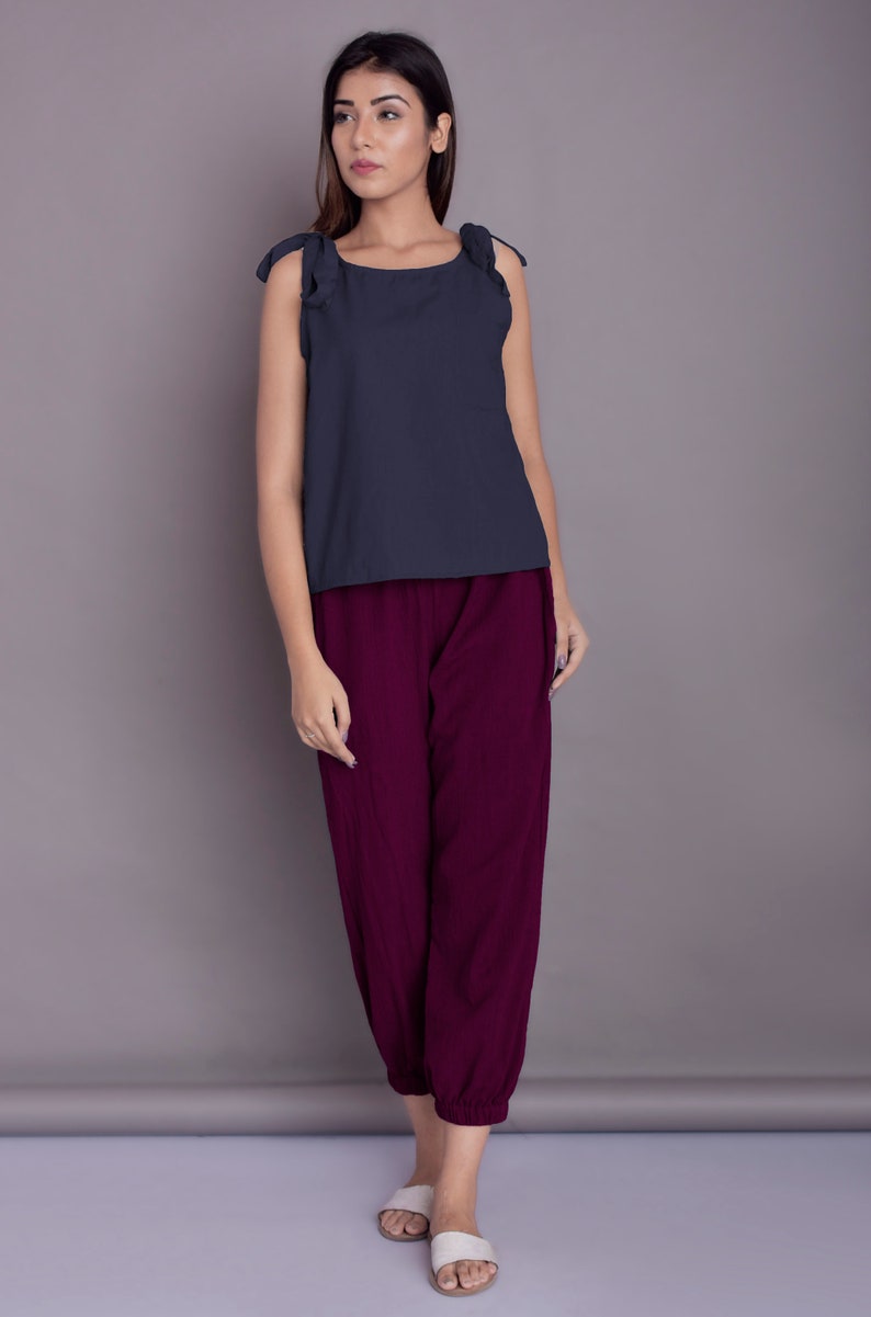 May include: A woman is wearing a dark blue sleeveless top with ruffled straps and burgundy trousers. She is standing on a light grey surface.
