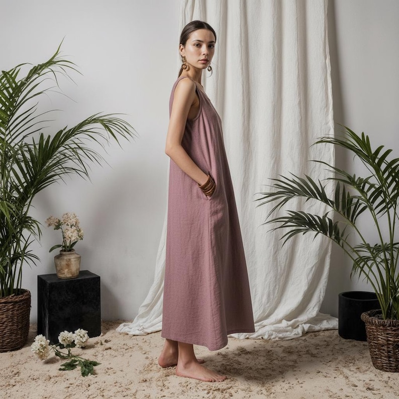 Op de afbeelding: Een mouwloze, stoffig roze maxi-jurk met een V-hals en zakken. De jurk is gemaakt van een zachte, vloeiende stof. Het model draagt houten oorringen en een bruine armband. De achtergrond omvat planten en een wit gordijn.