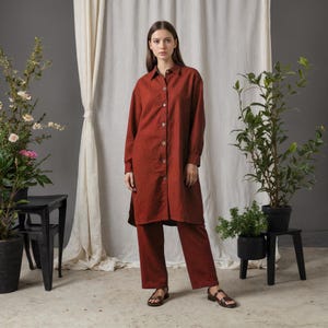 May include: A rust-colored linen shirt dress and matching pants. The long-sleeved shirt dress has a button-down front and a collared neckline. The pants are straight-legged. The outfit is paired with brown sandals. The background includes plants and a neutral-colored curtain.