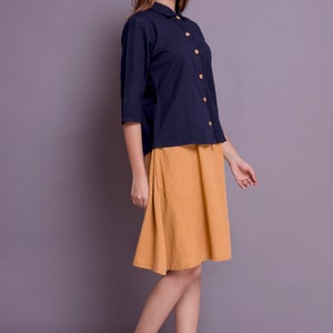 Pu&ograve; includere: Una donna indossa una camicia blu navy con colletto e bottoni in legno e una gonna giallo senape. Indossa sandali beige con zeppa.