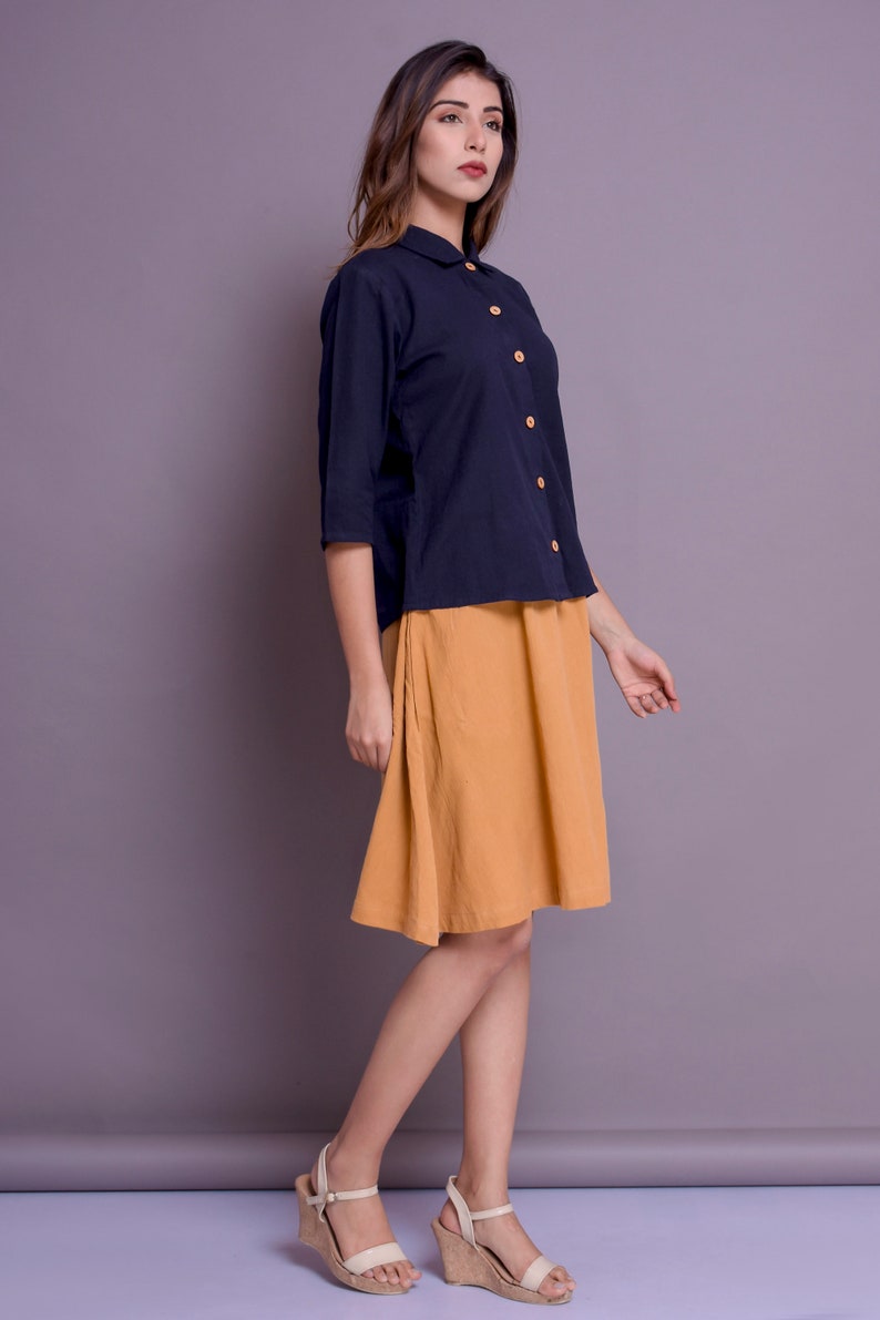 Pu&ograve; includere: Una donna indossa una camicia blu navy con colletto e bottoni in legno e una gonna giallo senape. Indossa sandali beige con zeppa.