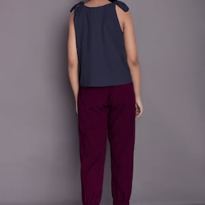 May include: A woman is wearing a dark blue sleeveless top with tie straps and maroon trousers. The trousers have a cuffed hem.
