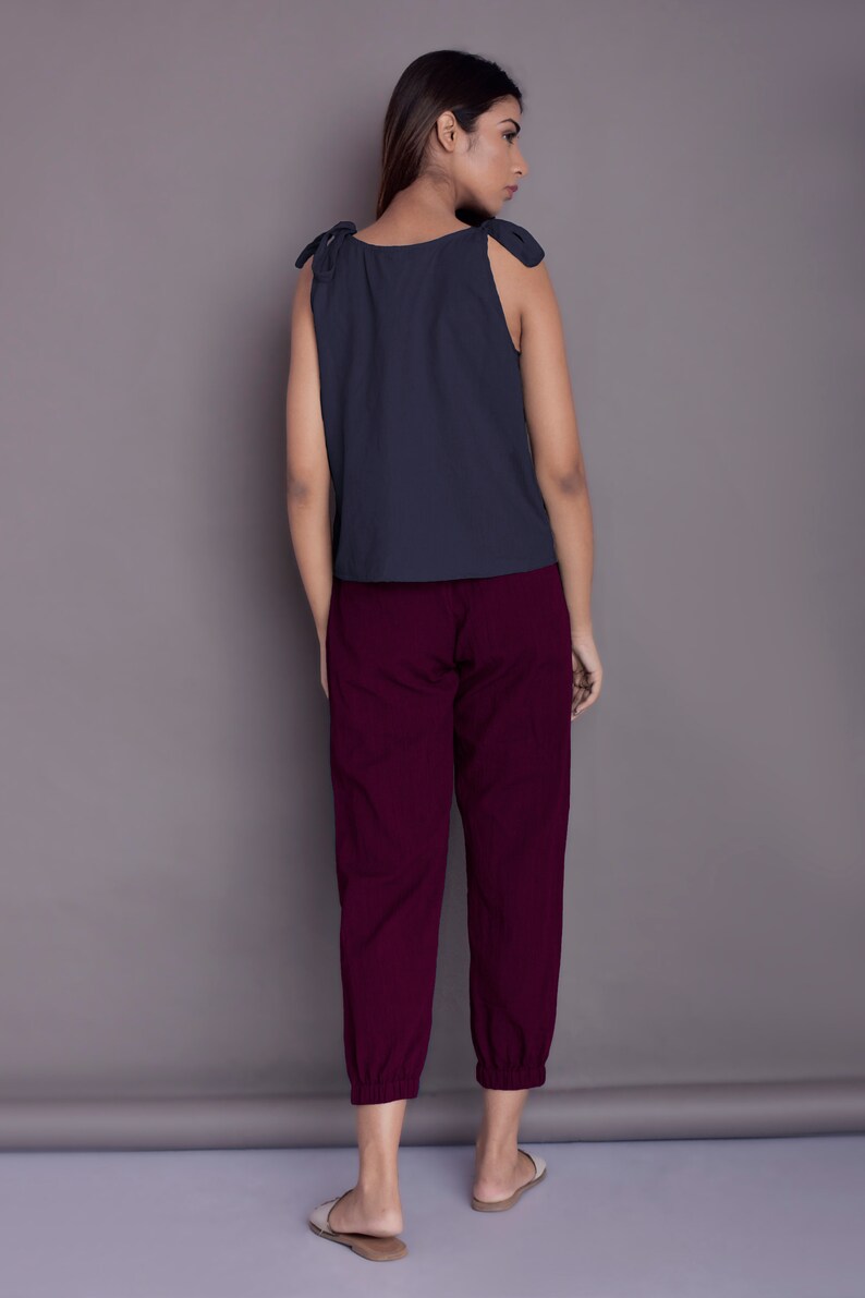 May include: A woman is wearing a dark blue sleeveless top with tie straps and maroon trousers. The trousers have a cuffed hem.
