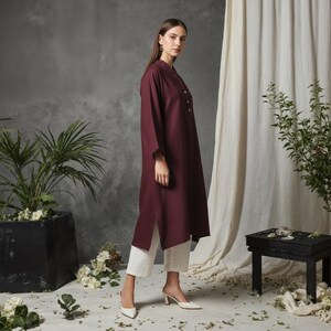 May include: A long-sleeved, burgundy tunic dress with a mandarin collar and button details. The dress is paired with white cropped pants and white heeled mules. The outfit is styled in a studio setting with plants and a neutral backdrop.