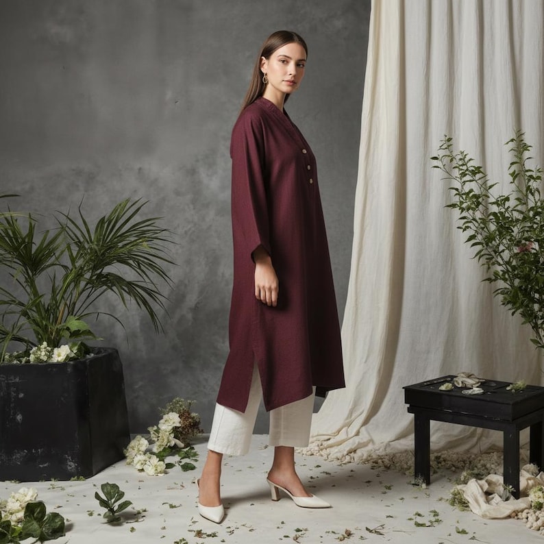 May include: A long-sleeved, burgundy tunic dress with a mandarin collar and button details. The dress is paired with white cropped pants and white heeled mules. The outfit is styled in a studio setting with plants and a neutral backdrop.