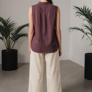 May include: A sleeveless, plum-colored top and cream-colored wide-leg pants. The top has a button-down collar. The pants are paired with black sandals. The outfit is displayed in a minimalist setting with potted plants.