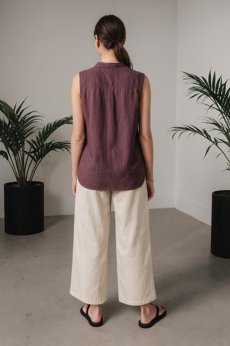 May include: A sleeveless, plum-colored top and cream-colored wide-leg pants. The top has a button-down collar. The pants are paired with black sandals. The outfit is displayed in a minimalist setting with potted plants.