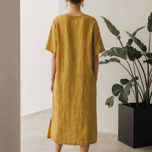 May include: Mustard yellow linen dress with short sleeves and a knee-length hem. The dress has a simple, relaxed fit and a side slit. The model is wearing black heeled sandals. A large potted plant is in the background.