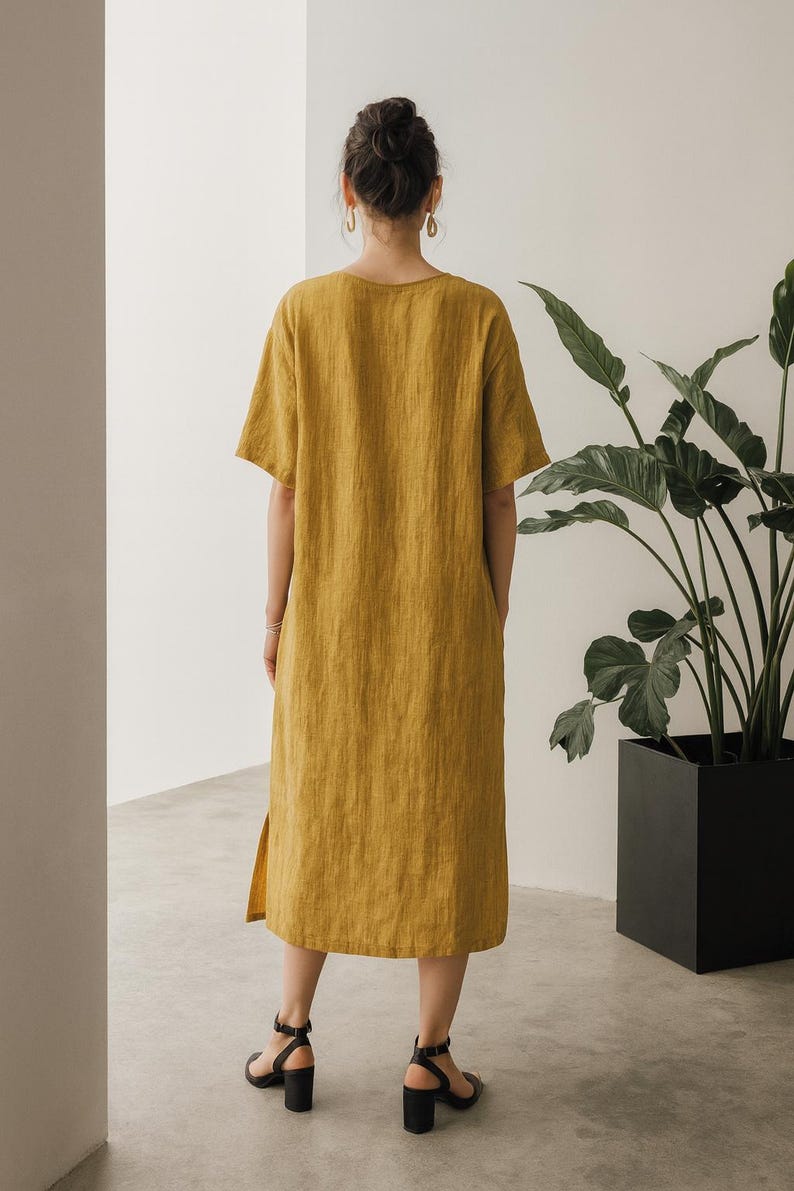 May include: Mustard yellow linen dress with short sleeves and a knee-length hem. The dress has a simple, relaxed fit and a side slit. The model is wearing black heeled sandals. A large potted plant is in the background.