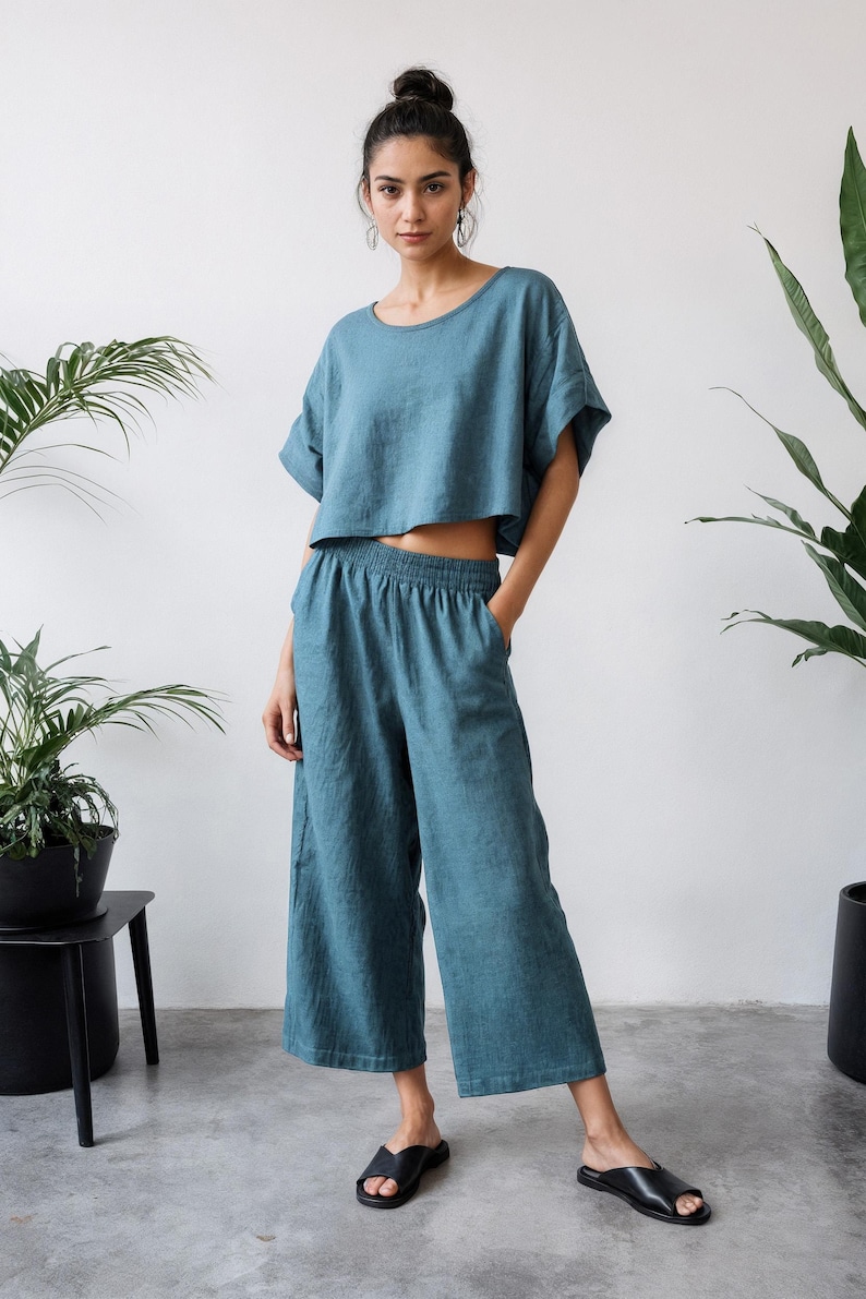 May include: A teal two-piece linen outfit. The set includes a short-sleeved crop top and wide-leg pants with an elastic waistband. The model is wearing black sandals. The outfit is displayed in a bright, minimalist setting with potted plants.