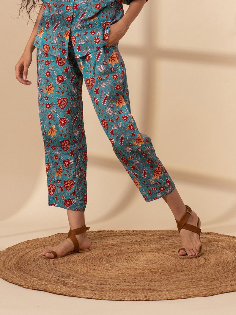 May include: Teal blue trousers with a floral print in shades of red, orange, and white. The trousers are cropped and have a wide leg.