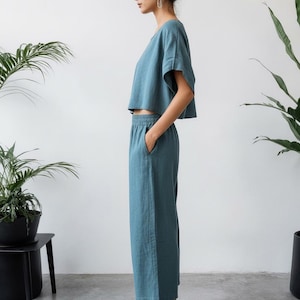 May include: A teal two-piece outfit, consisting of a short-sleeved crop top and wide-leg pants. The outfit is made of a soft, flowing fabric. The person is wearing black sandals. The background is a plain white wall with potted plants.
