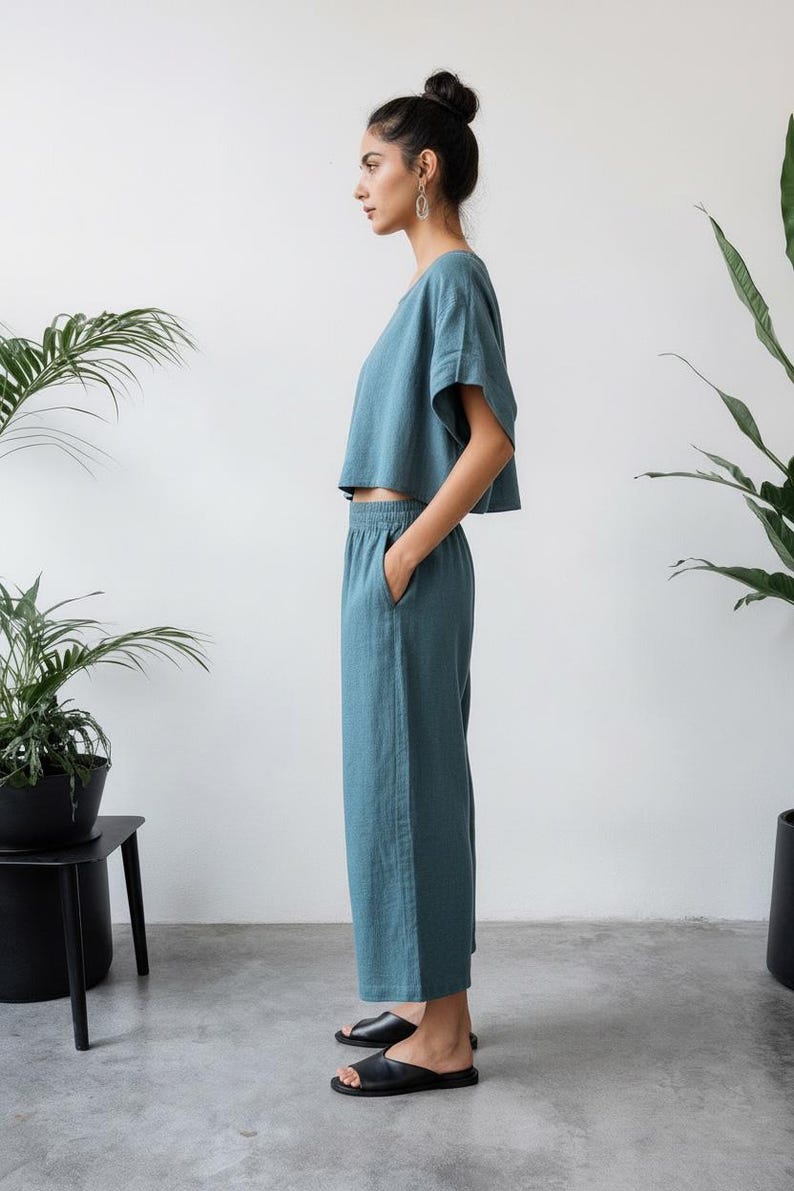 May include: A teal two-piece outfit, consisting of a short-sleeved crop top and wide-leg pants. The outfit is made of a soft, flowing fabric. The person is wearing black sandals. The background is a plain white wall with potted plants.