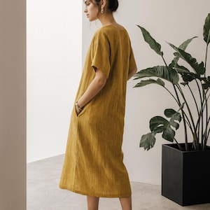 May include: Mustard yellow linen dress with short sleeves and pockets. The dress is knee-length and paired with black heeled sandals. A large potted plant is in the background.
