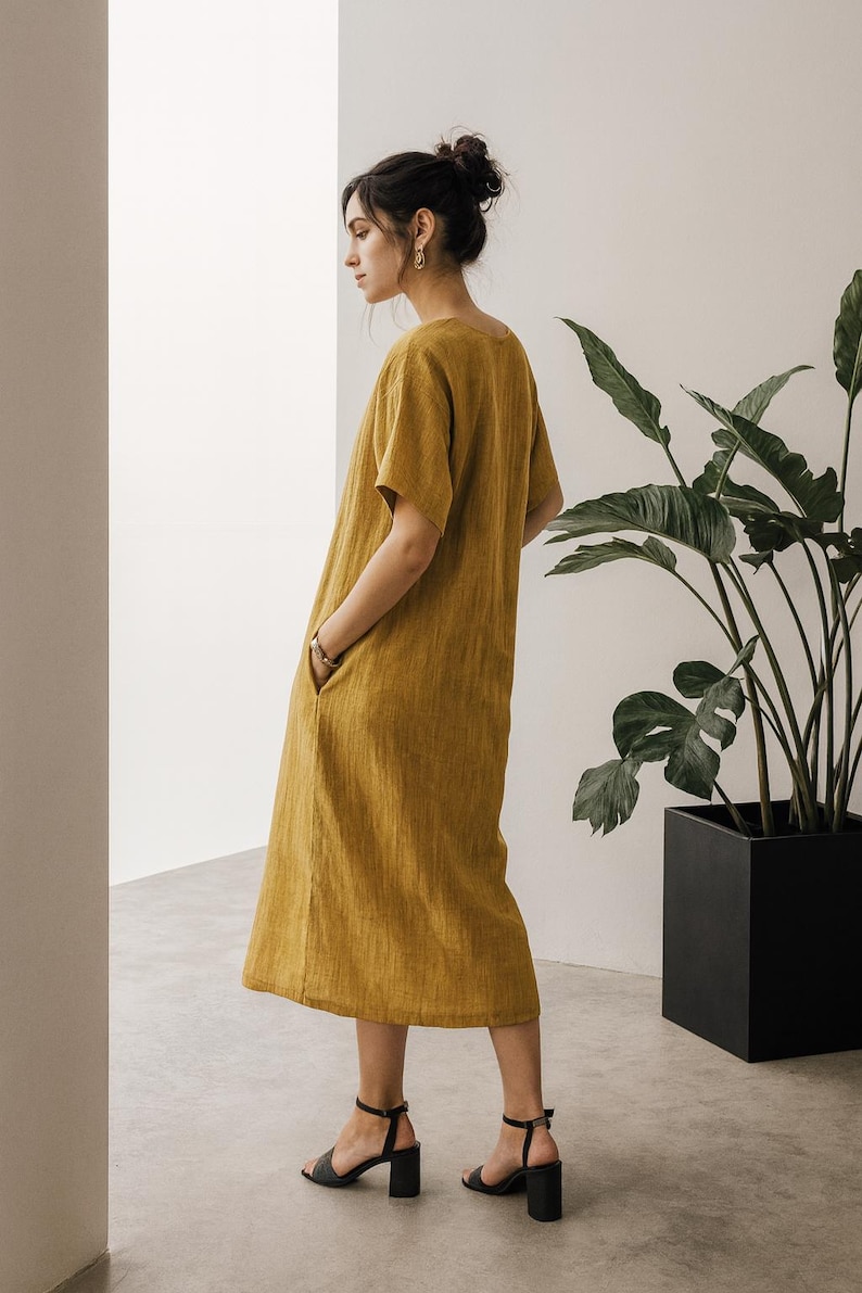 May include: Mustard yellow linen dress with short sleeves and pockets. The dress is knee-length and paired with black heeled sandals. A large potted plant is in the background.