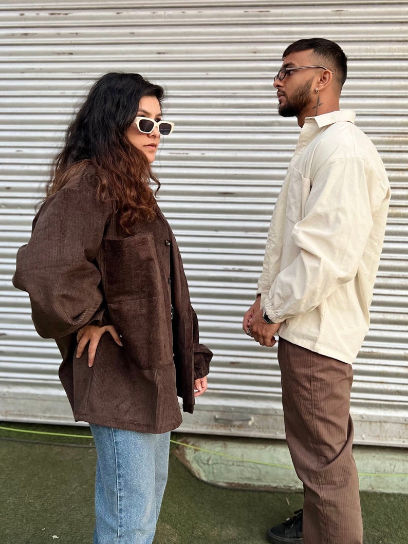 Puede incluir: Una persona lleva una camisa de pana marr&oacute;n con vaqueros azules y gafas de sol blancas, junto a otra persona con una camisa color crema y pantalones de pana marrones. El fondo es una persiana met&aacute;lica gris.