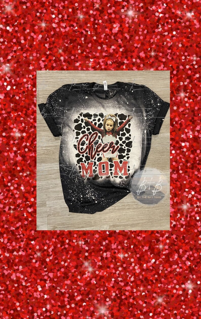 May include: Black t-shirt with a bleached design and a graphic of a young cheerleader in a red and white uniform. The words "Cheer MOM" are printed in red and white script.
