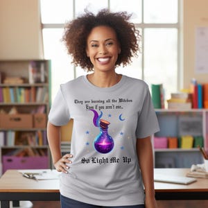 May include: A light gray t-shirt with the text "They are burning all the Witches Even if you aren't one... So Light Me Up." The shirt features a purple potion bottle design with stars and a crescent moon.