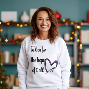 May include: White sweatshirt with the text "To live for the hope of it all" and a purple heart design. The word "hope" is written inside the heart. The sweatshirt is a crew neck style.