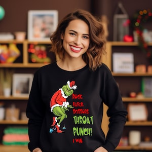 Może przedstawiać: Czarna bluza z nadrukiem Grincha w stroju Świętego Mikołaja. Grinch jest zielony i ma na sobie czerwono-biały strój. Tekst brzmi "Rock, Paper, Scissors, Throat Punch! I Win." Tekst jest w kolorze zielonym i czerwonym.