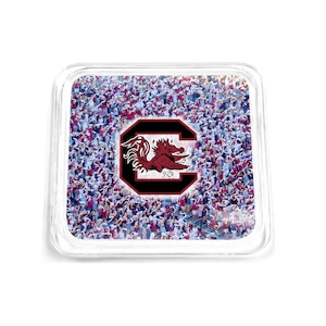 May include: A clear glass coaster with a red and black University of South Carolina Gamecocks logo on a background of a crowd of people wearing red and white.