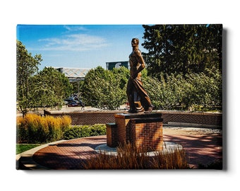May include: A bronze statue of a man in a walking pose, standing on a brick pedestal. The statue is surrounded by green trees and bushes. The statue is in front of a brick building with a green sign.