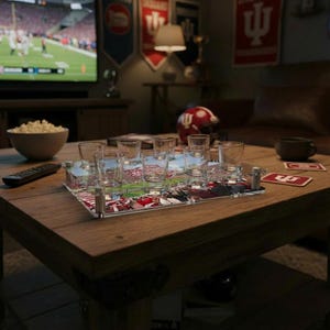 May include: A wooden coffee table displays a clear shot glass serving tray with six shot glasses. The tray has a sports-themed design. A bowl of popcorn, a remote, and playing cards are also on the table. A television shows a football game.