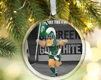 May include: A round clear ornament featuring a graphic of a Spartan mascot in green and white armor holding a flag. The background displays the text GO GREEN GO WHITE in bold letters. The ornament hangs from a white ribbon loop for display.