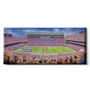 May include: A panoramic view of a packed football stadium with a green field and a large crowd of fans wearing blue and orange. The stadium is surrounded by a large number of seats and a scoreboard displaying the word "Gator."
