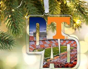 May include: A clear acrylic holiday ornament shaped like the letters DAD, featuring a photographic print of a football stadium filled with fans wearing orange. The letter T is printed in solid orange at the top, and a white ribbon is attached.