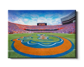 Op de afbeelding: Een panoramisch uitzicht op een volgepakt voetbalstadion met een groen veld en een groot oranje en blauw Gator-koplogo in het midden. Het scorebord geeft "GATORS" aan.